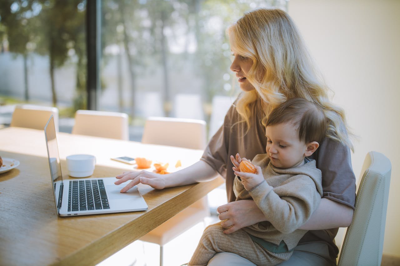 about-us A mother works on a laptop at home while holding her baby, showcasing remote working and parenting.